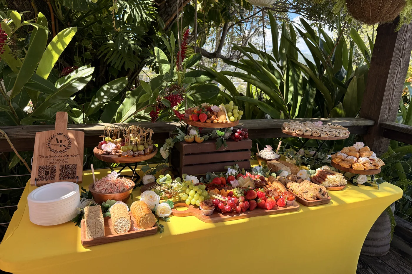 Mesa de grazing al aire libre en un jardín tropical, con el board de A&K y una gran variedad de charcutería y frutas